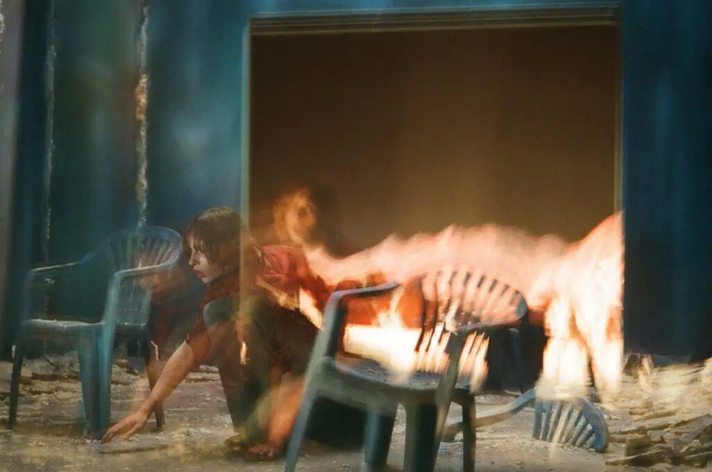 An artistic, long-exposure shot of Lauren Auder in a dark setting with motion blur and flames, featuring plastic chairs and a surreal, high-contrast aesthetic.