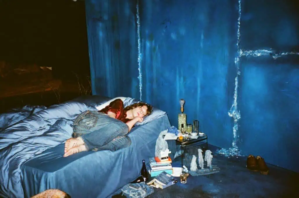 Lauren Auder lying on a blue bed in a room with matching blue walls, surrounded by books and bottles, creating a moody and cinematic atmosphere.