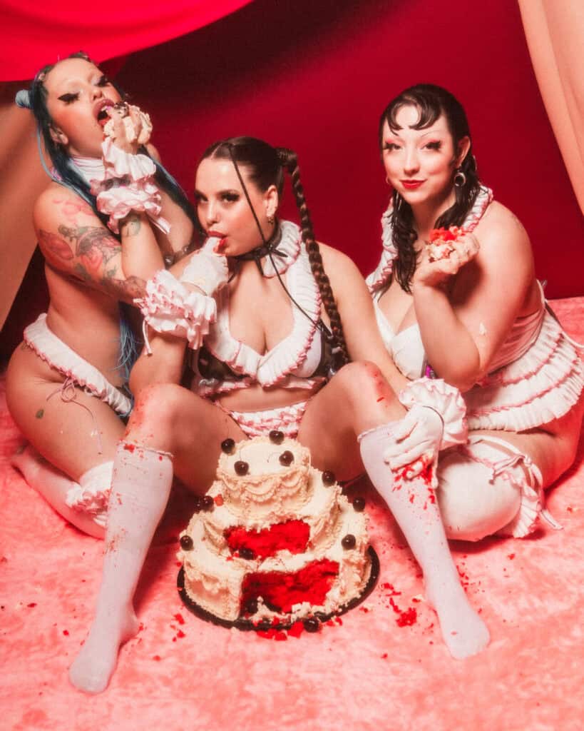 Three women in matching frilly lingerie and white thigh-high socks sit on a pink floor, engaging in playful cake sitting as they eat cream-covered cake with their hands. The red, draped fabric backdrop adds to the theatrical atmosphere.
