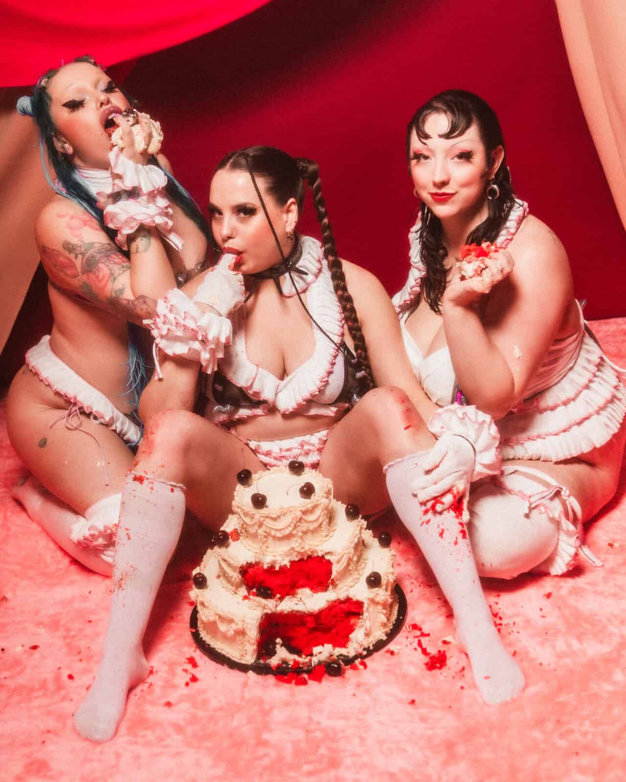 Three women in matching frilly lingerie and white thigh-high socks sit on a pink floor, engaging in playful cake sitting as they eat cream-covered cake with their hands. The red, draped fabric backdrop adds to the theatrical atmosphere.