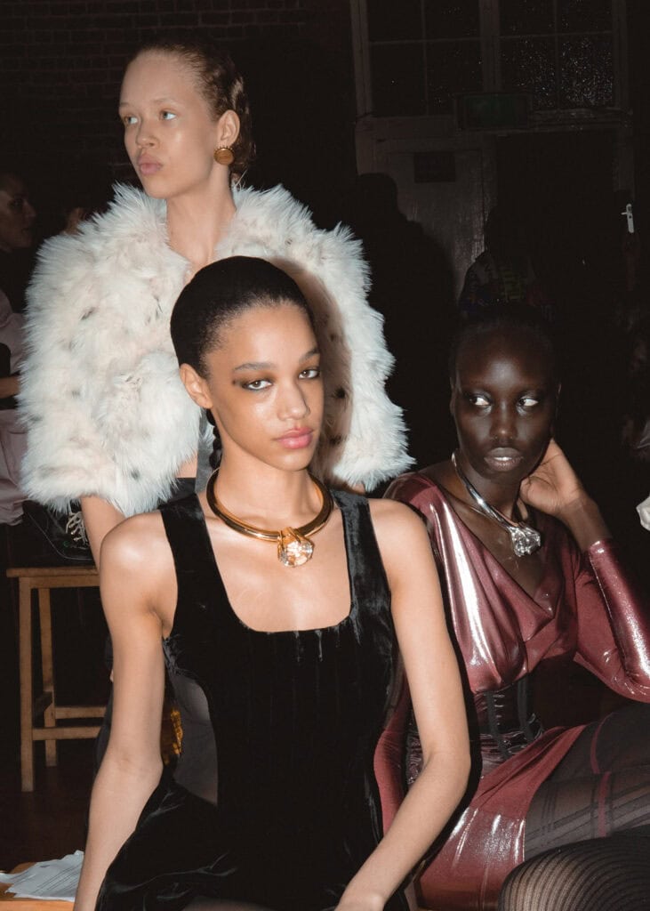 Three fashion models pose indoors: one in a white faux fur jacket, one in a black velvet dress with a gold necklace, and one in a shiny maroon outfit with dramatic makeup. The setting appears backstage or at a fashion event.