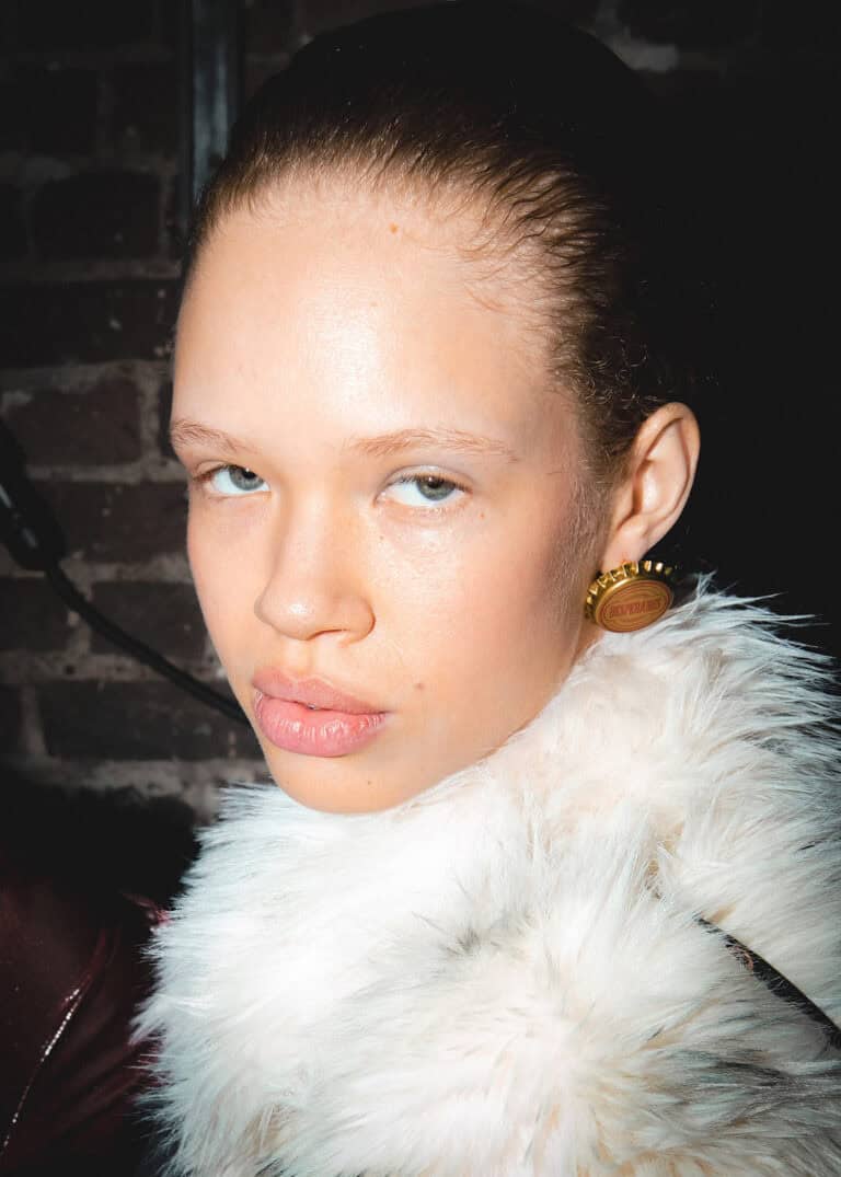 A person with light skin, blue eyes, and slicked-back hair wears a white Sinead Gorey faux fur garment and gold circular earrings. They pose against a brick wall, looking directly at the camera with a serious expression.