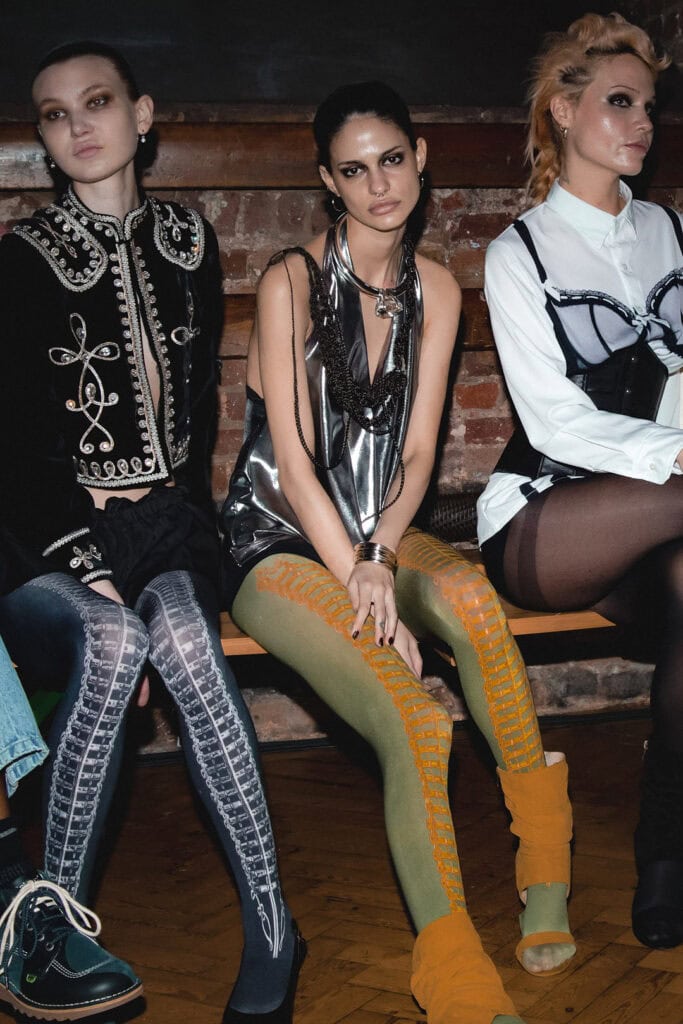Three women sit side by side on a bench, dressed in eclectic, Sinead Gorey fashionable outfits with bold patterns, metallic fabrics, and statement tights, against a rustic brick wall background.