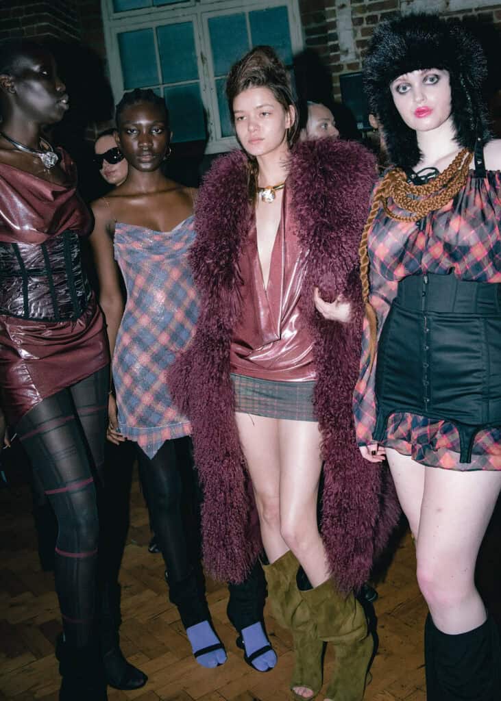 A group of five models in bold, eclectic outfits pose indoors. They wear plaid patterns, sheer fabrics, chunky accessories, and statement outerwear, with striking makeup and varied hairstyles, standing on a wooden floor under bright lighting.