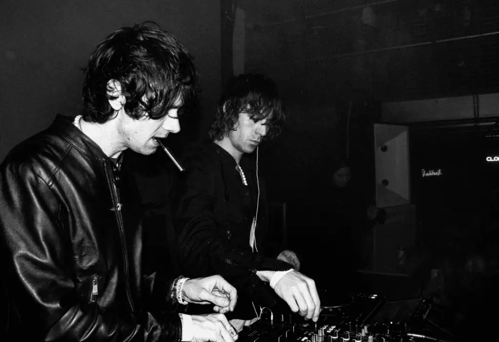 Black and white candid shot of The Hellp duo performing a DJ set in a dark club, with one member wearing a leather jacket and smoking.