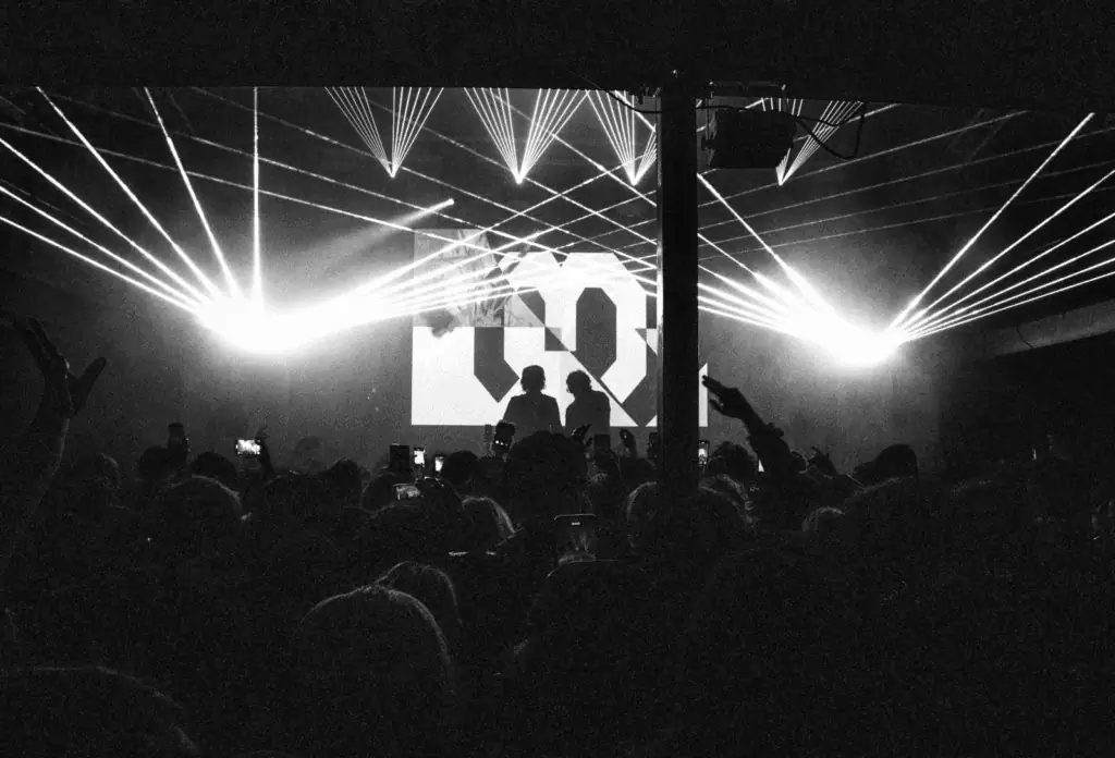 The Hellp performing live on stage silhouetted against intense white laser beams and a crowded audience at a high-energy concert.