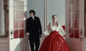 A man in a black suit and a woman in a dramatic red ball gown with sheer white sleeves stand side by side in an ornate room, framed by open white doors—a still from "Wuthering Heights" movie.