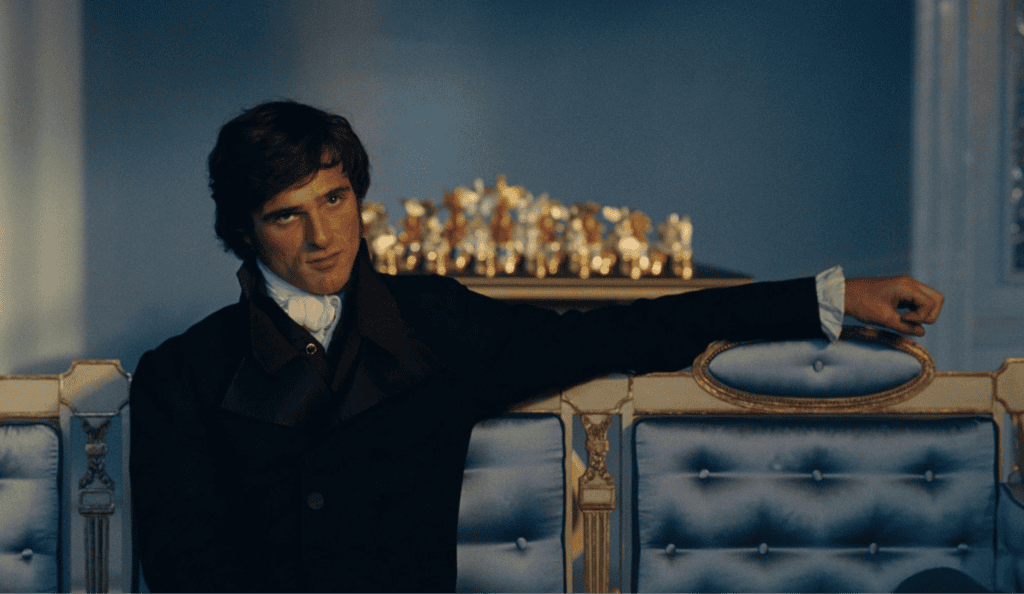 A young man in a dark suit sits casually on a blue, ornate sofa with tufted upholstery, his arm resting on the back. In the background, there is a golden chess set on a table.