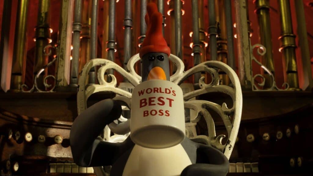 A clay penguin with a red hat sits on an ornate chair, holding a white mug labeled WORLDS BEST BOSS in front of a decorated pipe organ.