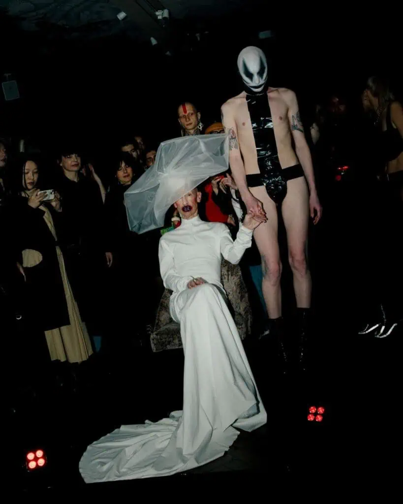 A dramatic performance by Dahc Dermur VIII, featuring a person in a long white gown and veil holding hands with a performer in a white mask and black fetish-inspired attire.