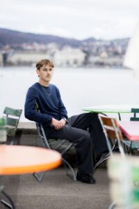 A portrait of author Nelio Biedermann sitting at an outdoor café table with a blurred city and lake background, wearing a dark blue sweater and grey trousers.