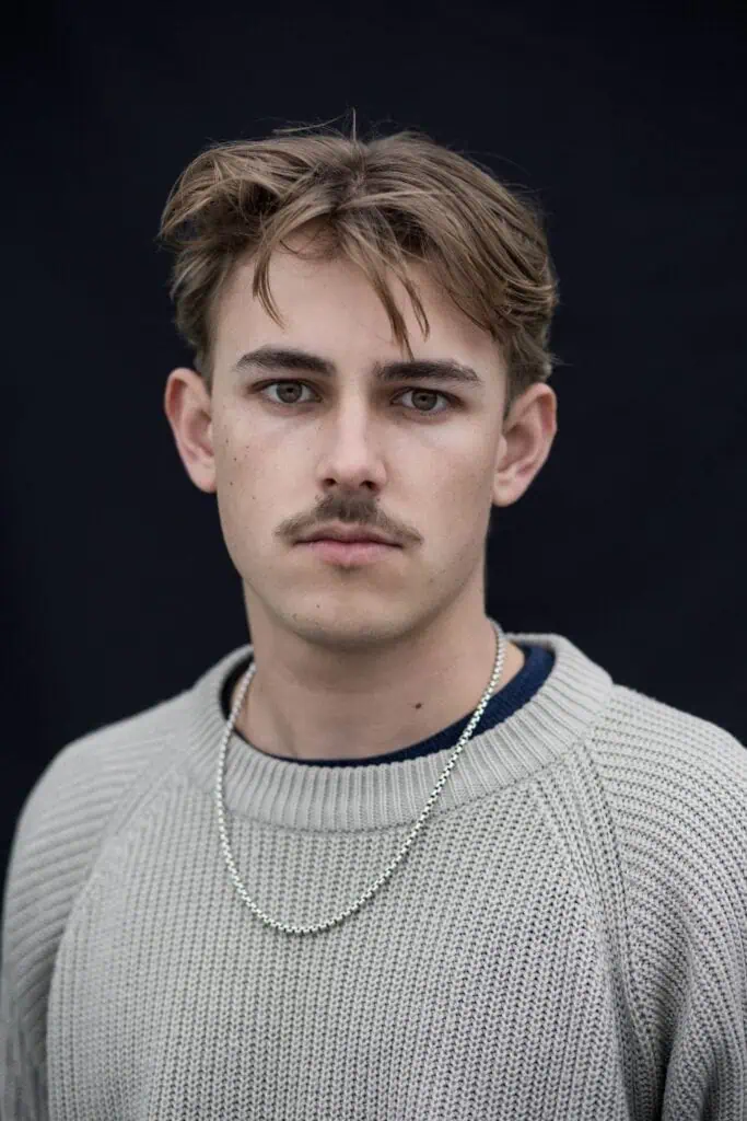 A close-up studio portrait of Nelio Biedermann looking directly at the camera, wearing a light grey textured sweater and a silver chain necklace against a dark background.