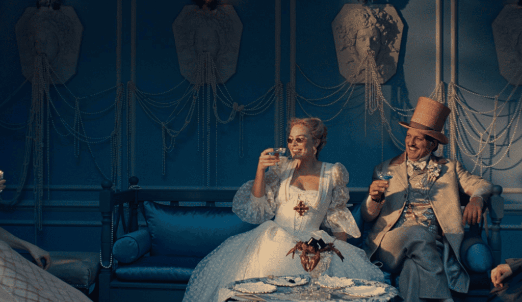 A woman in an elaborate white gown and a man in a gold suit and top hat sit on a blue sofa in an ornate room, raising glasses in a toast, with decorative wall art and pearls in the background.