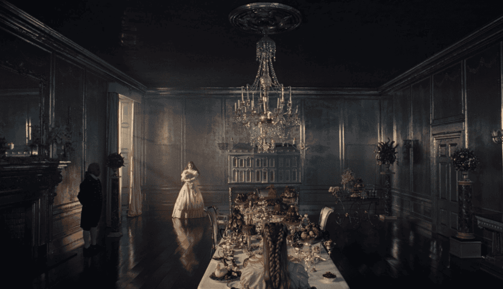 A lavish dining room with dark walls and a long table set with ornate dishes and fruit. A woman in a pink gown stands near the door while another person stands in the shadows. A large chandelier hangs from the ceiling.