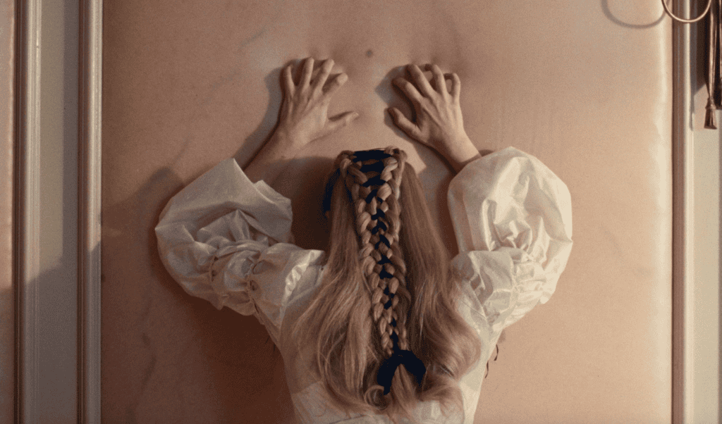 A person with long blond hair in a thick braid tied with blue ribbon presses their hands against a pale pink wall, wearing a white puffed-sleeve blouse. The scene is viewed from behind.