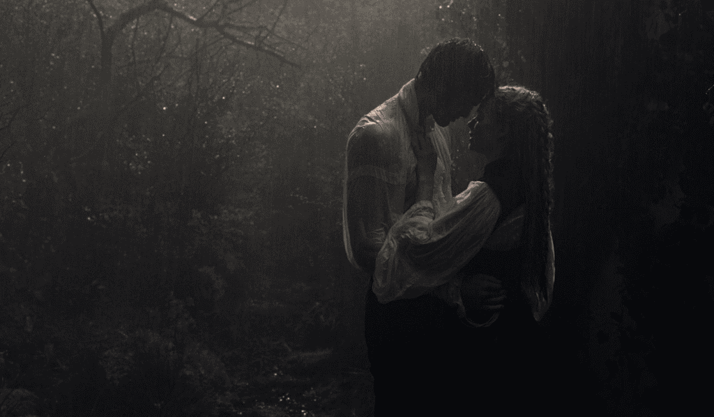 A man and woman stand closely together in a dim, misty forest, their foreheads touching as they embrace, both wearing old-fashioned white shirts and vests, with soft light filtering through the trees.