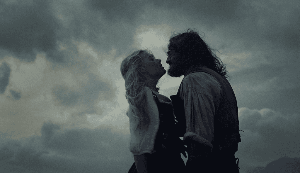 A woman and a man stand face to face outdoors under a dramatic, cloudy sky. The woman looks up at the man, both appearing intense and emotional in their rustic, historical clothing.