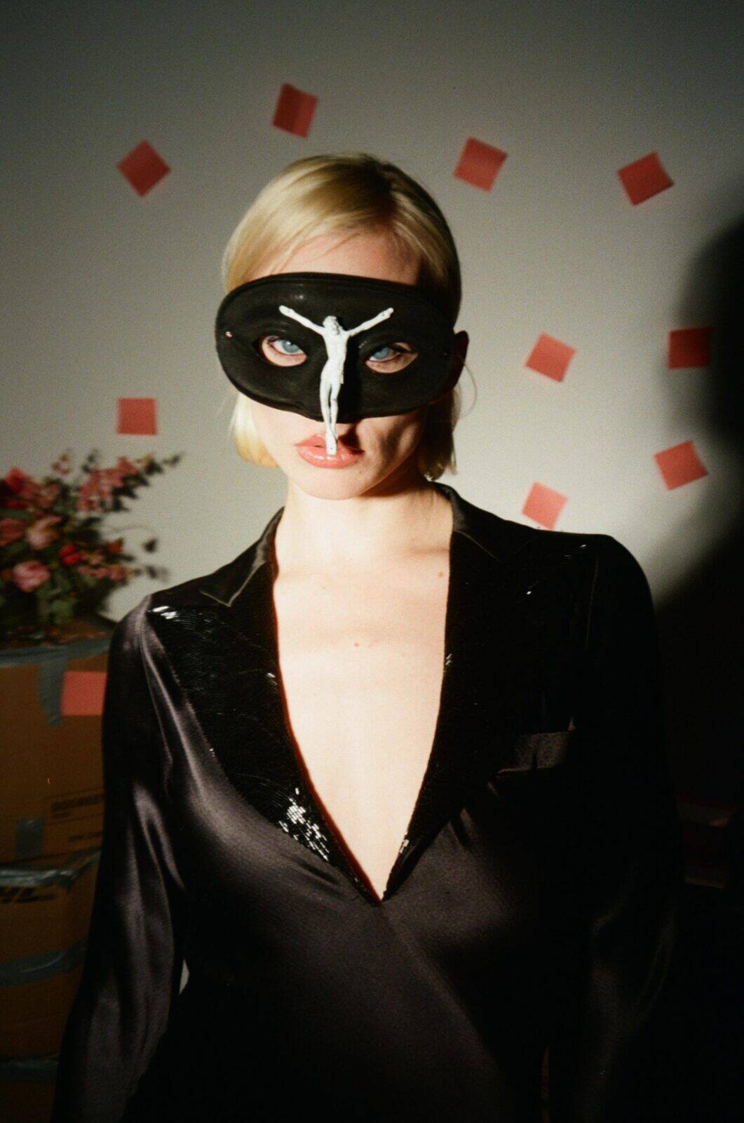 A person in a deep V-neck black dress wears a black mask with a small white figure attached. Red square decorations are on the wall behind, and a vase of flowers and cardboard boxes are partially visible.