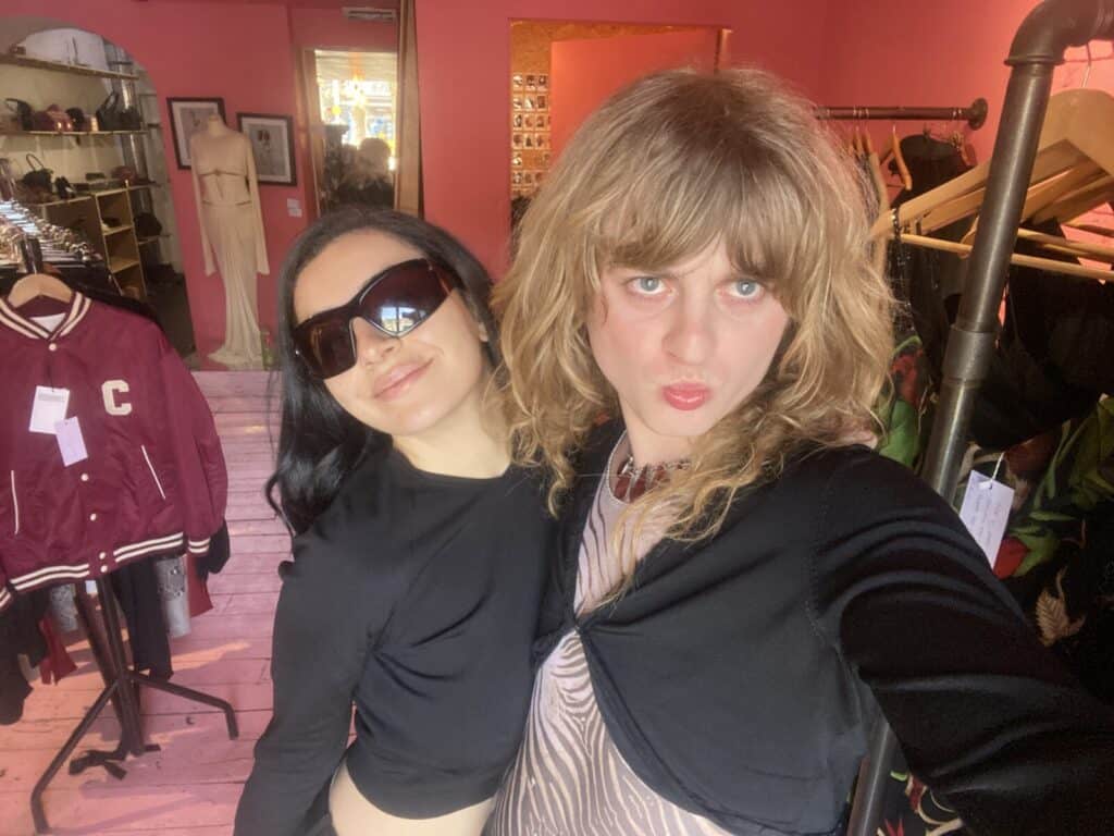 Two people pose for a selfie in a boutique with pink walls. One wears sunglasses and a black top; the other has long wavy blonde hair and a black jacket. Racks of clothes and a letterman jacket are visible in the background.