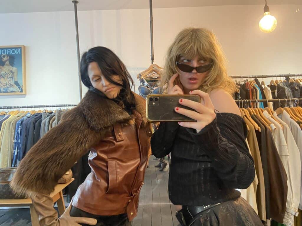 Two people pose stylishly in a clothing store. One wears a fur-collared brown jacket and gloves, while the other wears sunglasses and an off-the-shoulder black top, holding a phone to take a mirror selfie. Racks of clothes are in the background.