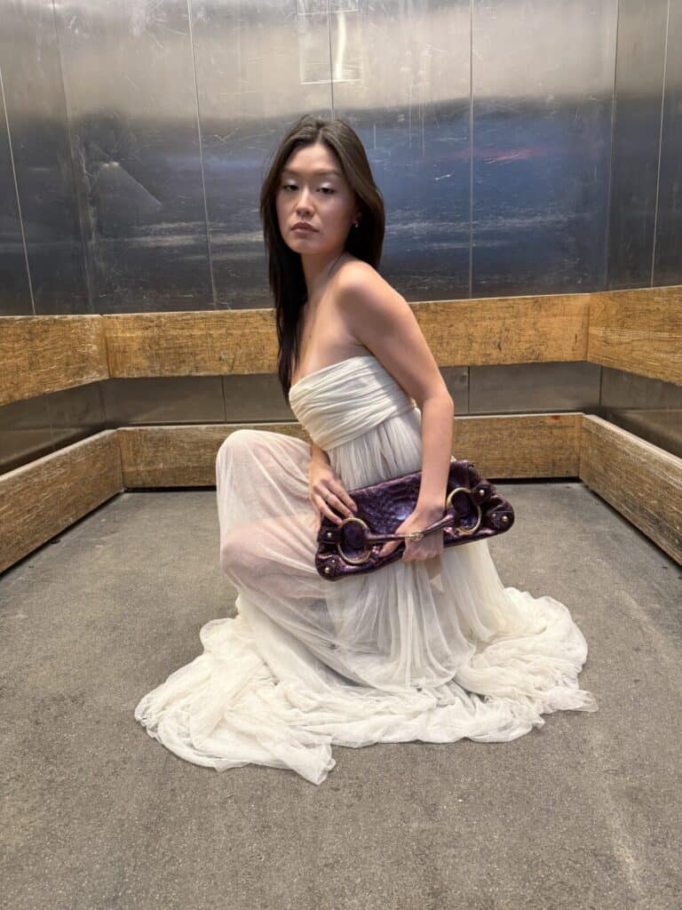 A woman in a strapless white gown kneels on the floor of a metallic, industrial-style room, holding a unique, sculptural purple clutch while looking over her shoulder with a calm expression.