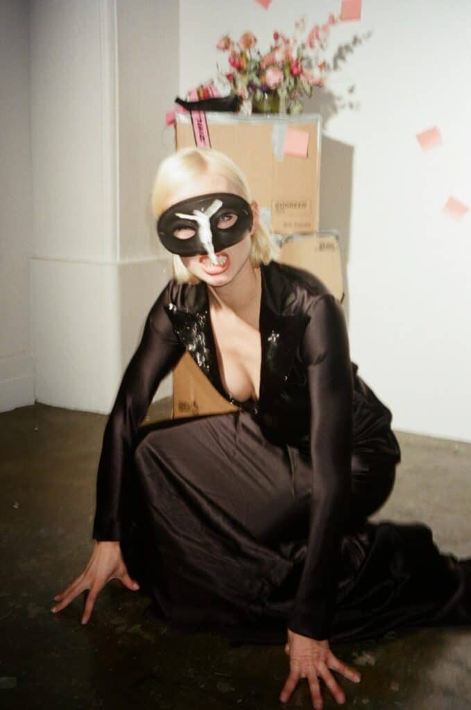 A person in a black mask and long black dress crouches on the floor in front of stacked cardboard boxes and a vase of flowers, with pink sticky notes on the wall behind them.