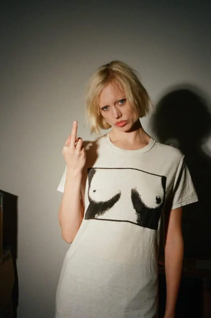 A woman with short blonde hair stands against a plain wall, wearing a white T-shirt with a printed graphic of breasts. She faces the camera and raises her left hand, giving the middle finger.