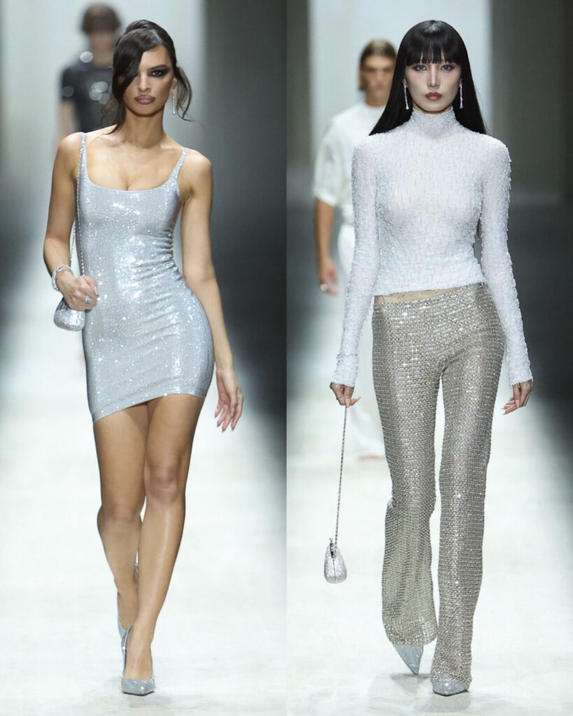 Two runway models showcase sparkly silver outfits; the left model wears a sleeveless mini dress, and the right model wears a long-sleeve turtleneck top with matching pants. Both carry small silver bags and wear silver heels.
