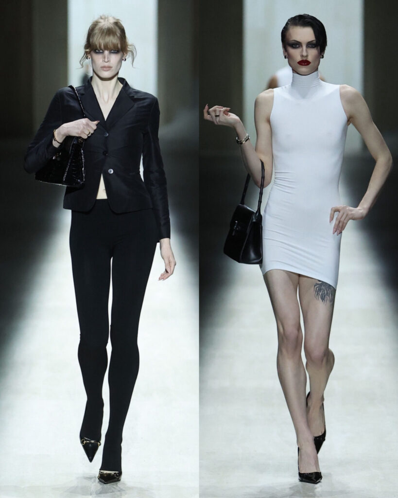 Two fashion models walk a runway; the left model wears a fitted black blazer, black leggings, and heels, while the right model wears a sleeveless white mini dress, black purse, heels, and displays a thigh tattoo.