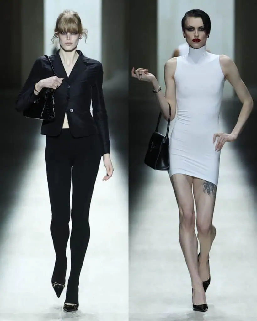 Two fashion models walk a Gucci AW26 runway; the left model wears a fitted black blazer, black leggings, and heels, while the right model wears a sleeveless white mini dress, black purse, heels, and displays a thigh tattoo.