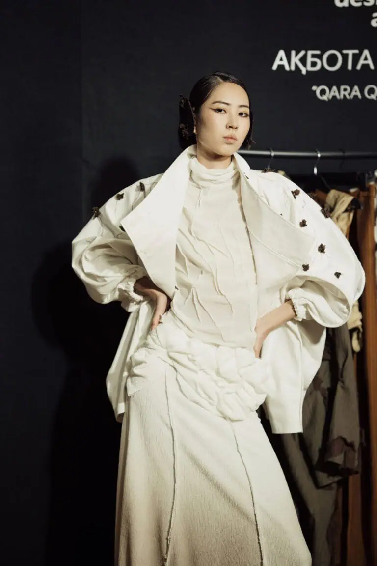 Model wearing Akbota Kapsalan’s white "Qara Qandyagash" collection, featuring a textured high-neck top and a jacket adorned with small dark floral accents at Visa Fashion Week Tashkent.