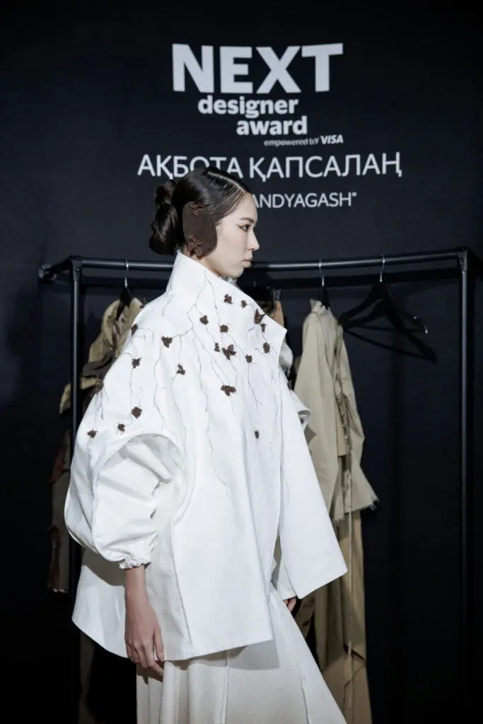 A side profile shot of a model wearing Akbota Kapsalan’s white "Qara Qandyagash" jacket, showcasing intricate brown floral branch embroidery and a matching felt ear accessory at the NEXT Designer Award in Tashkent.