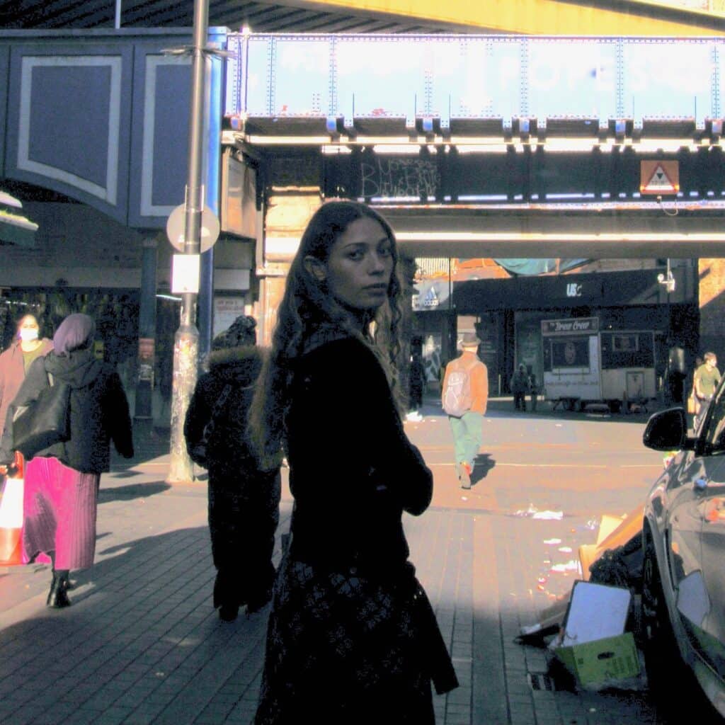 Alt-pop artist Alba Akvama standing in a sun-drenched urban street under a blue industrial bridge. A candid, cinematic shot reflecting a gritty, atmospheric aesthetic.