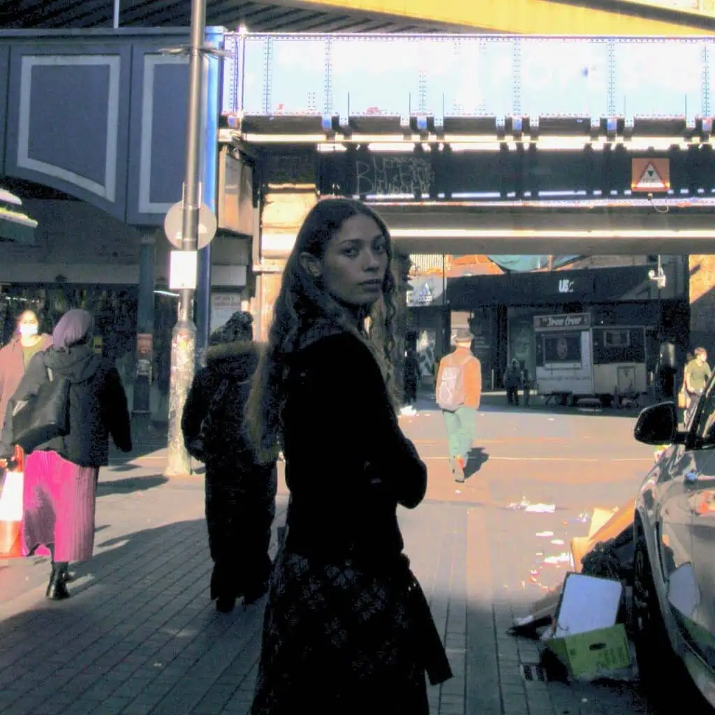 Alt-pop artist Alba Akvama standing in a sun-drenched urban street under a blue industrial bridge. A candid, cinematic shot reflecting a gritty, atmospheric aesthetic.