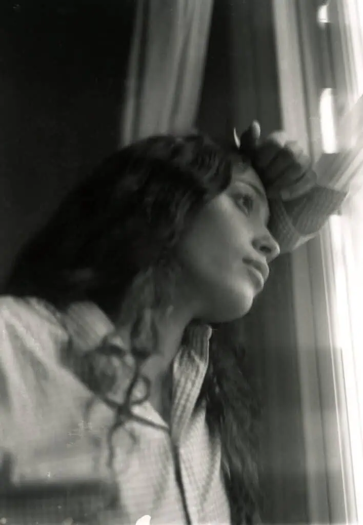 A grainy, black-and-white profile shot of Alba Akvama leaning against a window. Her hand is resting on her forehead in a contemplative pose as she gazes outside, creating a soft, cinematic mood.