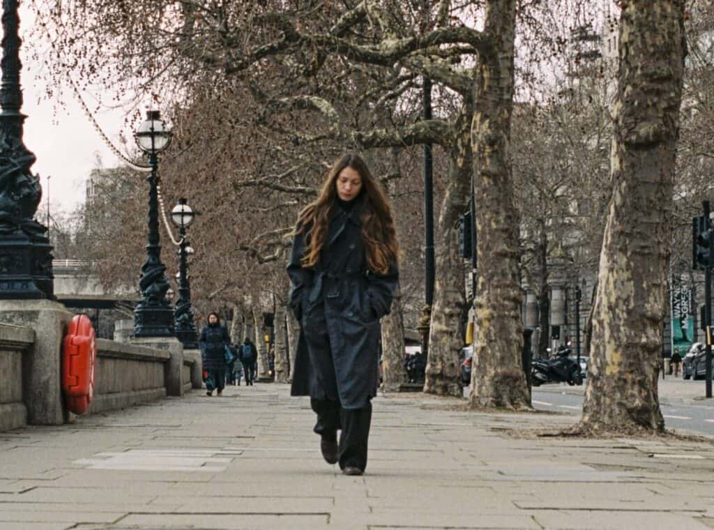 A wide shot of Alba Akvama walking down a tree-lined sidewalk in London. She is wearing a dark trench coat with her head slightly bowed, framed by bare winter trees and classic street lamps along the river.