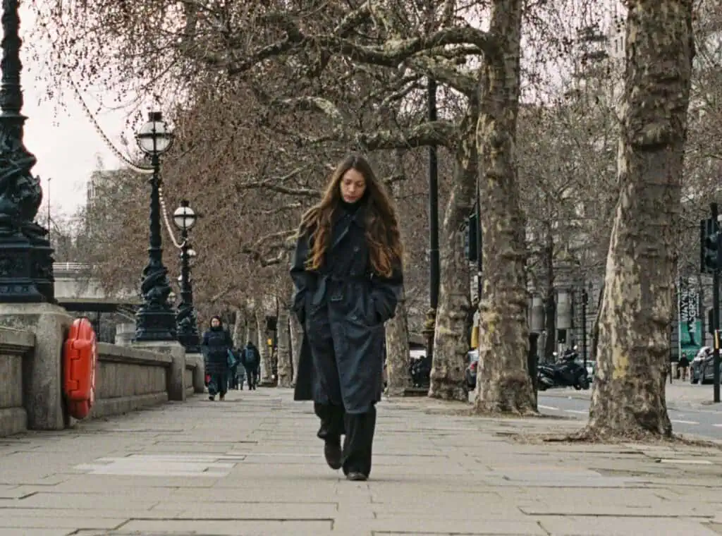 A wide shot of Alba Akvama walking down a tree-lined sidewalk in London. She is wearing a dark trench coat with her head slightly bowed, framed by bare winter trees and classic street lamps along the river.
