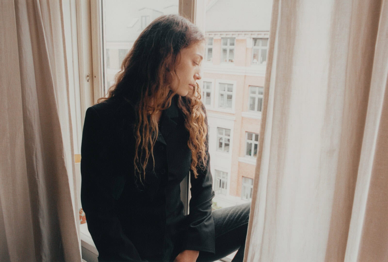 A medium shot of Alba Akvama sitting on a windowsill between soft, neutral curtains. She is wearing a black blazer and looking down pensively, with European-style brick architecture visible through the glass behind her.