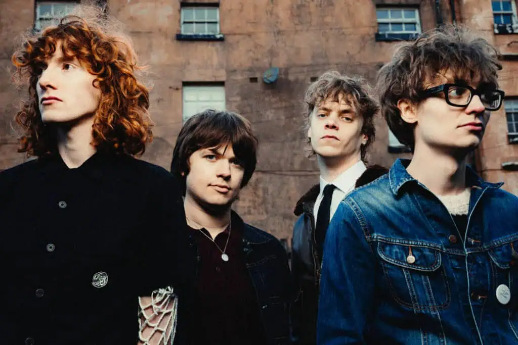 Portrait of the four members of The Sukis facing the camera against a weathered brick wall, including one with red curly hair in a black coat with mesh sleeves and another in a denim jacket with black-framed glasses — photograph by Nathan Cole