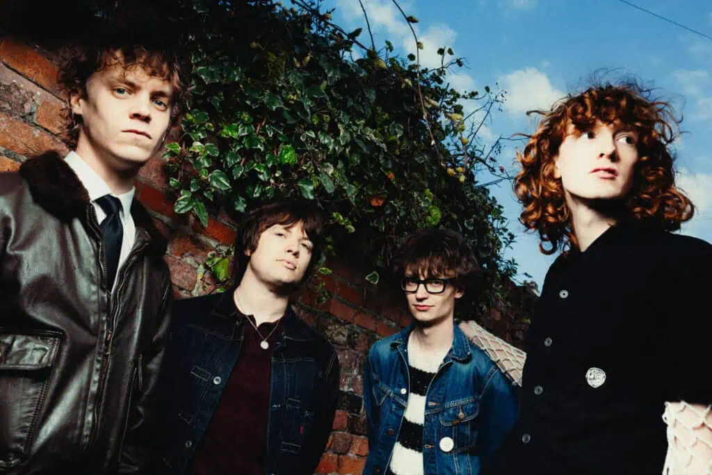 The Sukis photographed outdoors against an ivy-covered brick wall beneath a blue sky, one member in a leather jacket and tie, another with red curly hair looking upward — photograph by Nathan Cole
