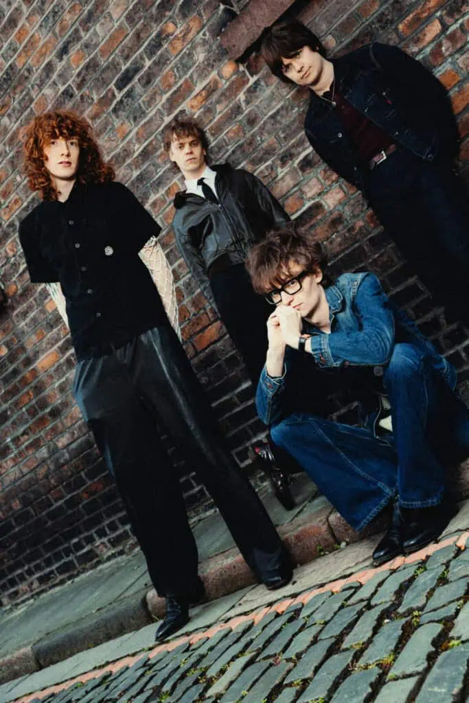 The four members of The Sukis posed in a brick alleyway, one crouching with hands clasped, another standing tall with red curly hair, captured in a moody editorial style — photograph by Nathan Cole