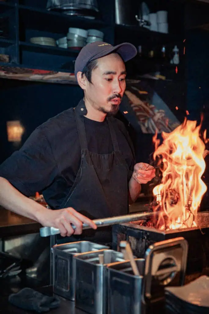 A chef at Yuzu Dining Bar in Amsterdam expertly grilling chicken over an open flame, capturing the authentic coal-fired yakitori cooking style.