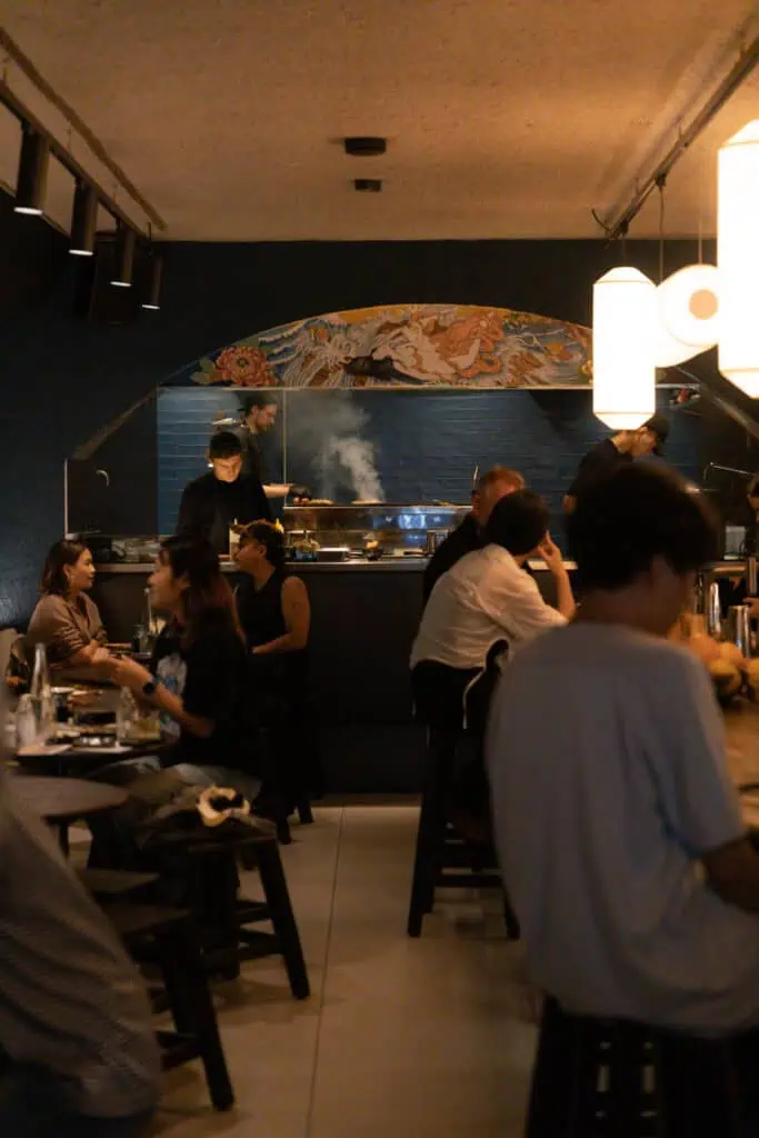 The moody, modern interior of Yuzu Dining Bar in Amsterdam, featuring a dark blue wall with Japanese art and customers dining near the open kitchen.