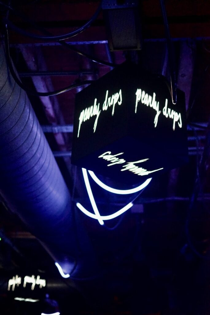 A glowing neon cube featuring the Pearly Drops logo hanging from the ceiling of a dark music venue.