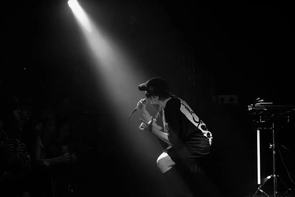A dramatic black and white shot of Pearly Drops performing on stage, with a single bright spotlight cutting through the dark venue.