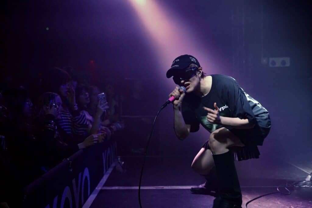Sandra Tervonen of Pearly Drops performing live in a purple spotlight, wearing a black cap and sunglasses at a concert.