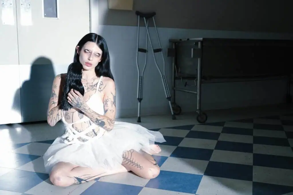 DeathbyRomy sitting on a blue checkered floor in a hospital setting, holding a long black hairpiece with crutches and a gurney blurred in the background.