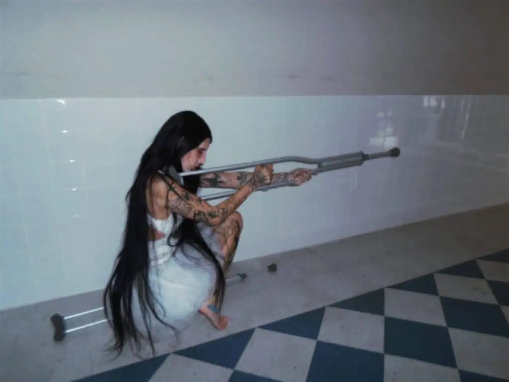 DeathbyRomy crouching in a white tiled hospital corridor, holding a crutch like a weapon as part of her "Art Against the World" editorial for The Cold Magazine.