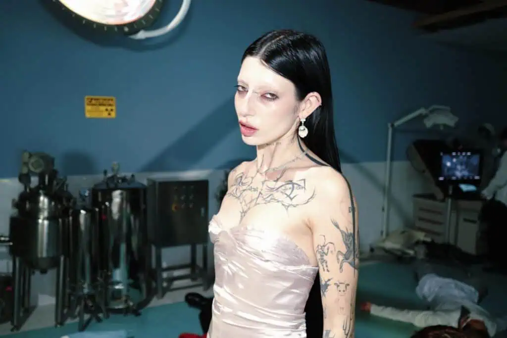 A high-fashion portrait of DeathbyRomy in a satin dress, posing in a clinical operating room with medical equipment and a body on the floor in the background.