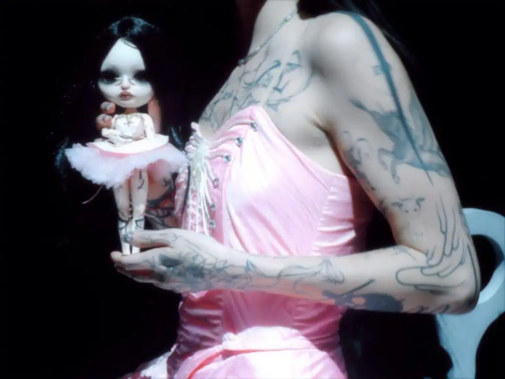 A close-up side profile of DeathbyRomy holding a small gothic doll that mirrors her own likeness, showcasing her intricate arm and chest tattoos.